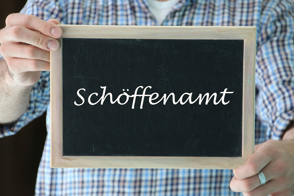 zwei Hände halten eine Schiefertafel mit der Aufschrift "Schöffenamt"