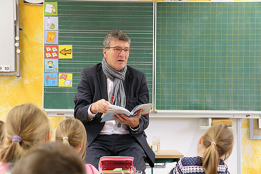 Minister Dieter Lauinger sitzt in einem Klassenraum vor der Tafel und liest den Schülern aus einem Buch vor