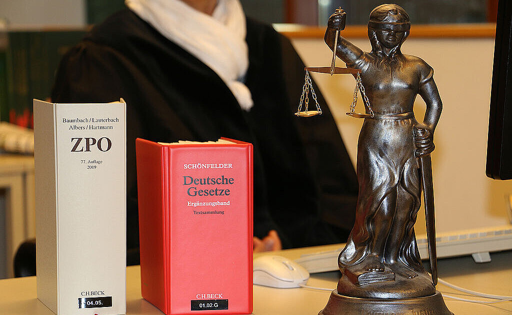 Ausschnitt von einer Richterin mit Robe vor einem PC und auf dem Tisch davor stehen Gesetzesbücher