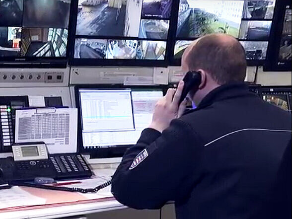 Ein Beamter des mittleren allgemeine Vollzugsdienstes sitzt in der Überwachungskanzel vor den Bildschirmen und telefoniert