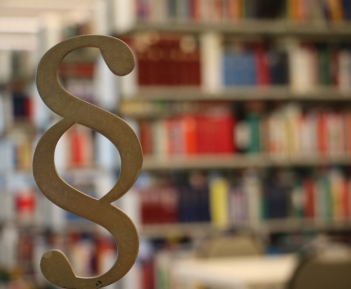 Paragraphenzeichen im Vordergrund vor Bücherregalen der Bibliothek Vor einem unscharf abgegbildeten Bücherregal befindet sich eine Kleinskulptur in Form eines Paragraphenhzeichens.