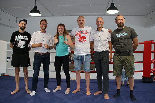Minister Dirk Adams sowie Mitglieder des La Familia Fightclub und der Hauptgeschäftsführer des Landessportbundes, Thomas Zirkel, stehen auf einer Trainingsmatte.