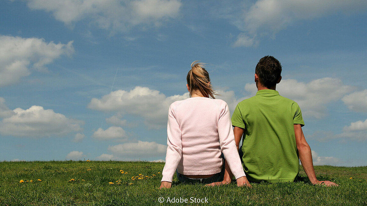 Ein junges Pärchen sitzt auf einer grünen Wiese mit Löwenzahn und schaut in den Himmel, der mit Schäfchenwolken bedeckt ist