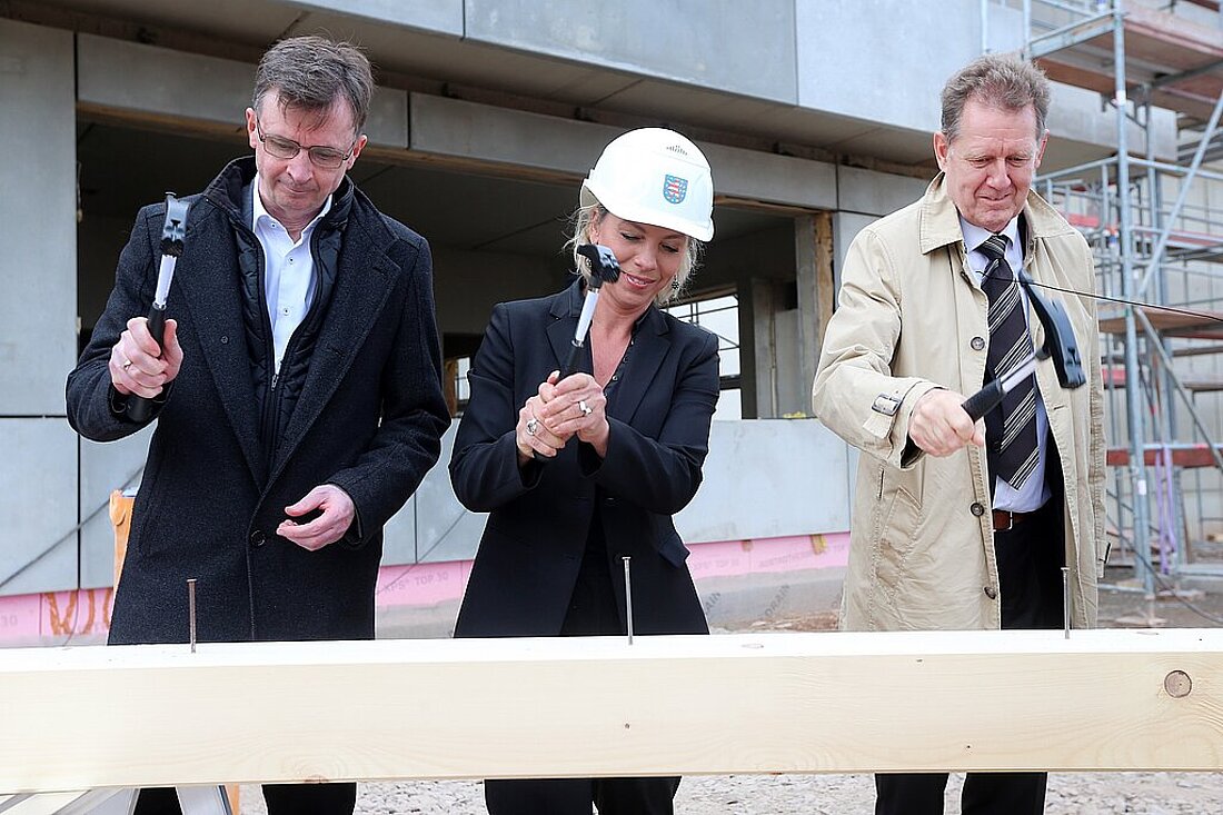 Richtfest JVA Zwickau-Marienthal Sebastian von Ammon, Staatssekretär im thüringischen Justizministerium, die thüringische Infrastrukturministerin Susanna Karawanskij und der Amtschef des Sächsischen Finanzministeriums Dirk Diedrichs klopfen einen Nagel in einen Balken.